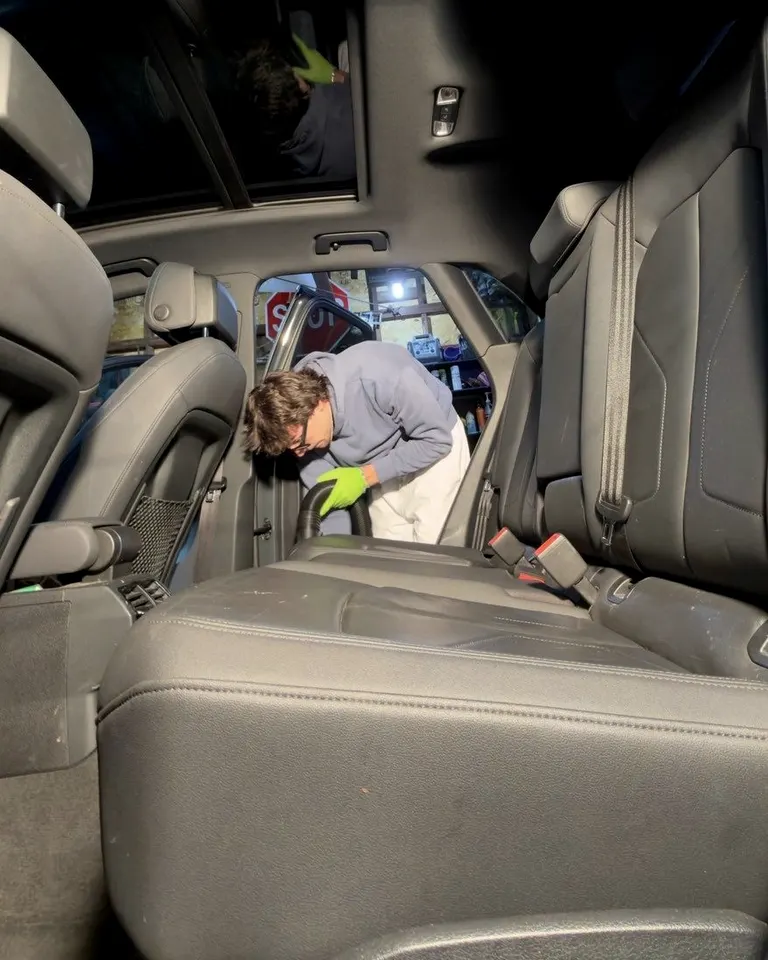Detailer in green gloves vacuuming the rear seats of an SUV through the open side door, seen from inside the cabin